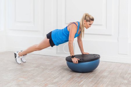 Photo for Full length portrait of concentrated woman in black shorts and blue top working in gym doing plank for abdominal muscles on bosu balance trainer, holding balance on fitness ball. Indoor studio shot. - Royalty Free Image