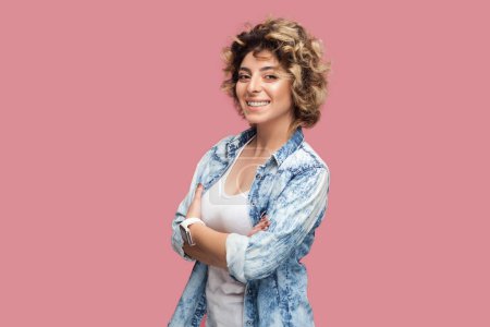 Photo for Portrait of confident happy smiling woman with curly hairstyle wearing blue shirt standing with crossed arms, looking at camera with positive expression. Indoor studio shot isolated on pink background - Royalty Free Image