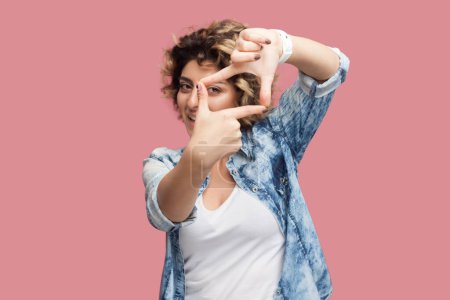 Téléchargez les photos : Portrait d'une femme photographe attirante concentrée avec une coiffure bouclée portant une chemise bleue regardant la caméra à travers le cadre des doigts. Studio intérieur tourné isolé sur fond rose. - en image libre de droit