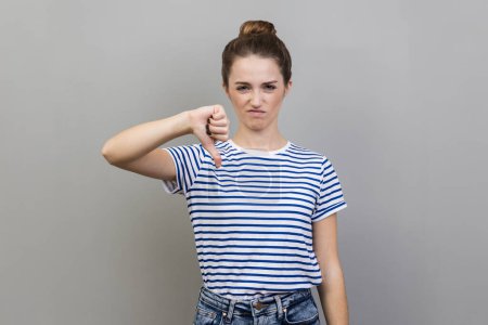 Photo for Portrait of unhappy unsatisfied young adult woman wearing striped T-shirt showing thumb down, dislike gesture, negative emotions, bad rating. Indoor studio shot isolated on gray background. - Royalty Free Image