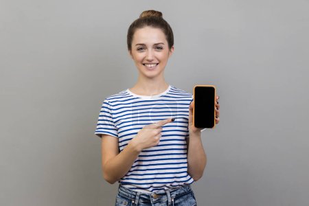 Photo for New mobile app. Portrait of woman wearing striped T-shirt pointing at cell phone and smiling at camera, showing telephone, copy space. Indoor studio shot isolated on gray background. - Royalty Free Image