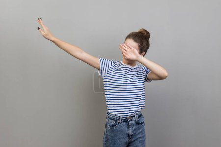Photo for Portrait of anonymous woman with bun hairstyle wearing striped T-shirt standing in dab dance pose, internet meme, celebrating success. Indoor studio shot isolated on gray background. - Royalty Free Image