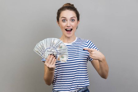 Photo for Portrait of surprised astonished woman wearing striped T-shirt pointing at fan of dollar banknotes, showing big money and looking at camera. Indoor studio shot isolated on gray background. - Royalty Free Image