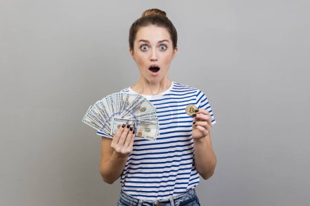 Photo for Portrait of surprised shocked woman wearing striped T-shirt holding gold bitcoin and fan of dollar bills, looking at camera with open mouth. Indoor studio shot isolated on gray background. - Royalty Free Image