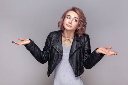 Photo for Portrait of puzzled confused woman with short hairstyle standing spreads hands aside, doesn't know what to do, wearing black leather jacket. Indoor studio shot isolated on grey background. - Royalty Free Image