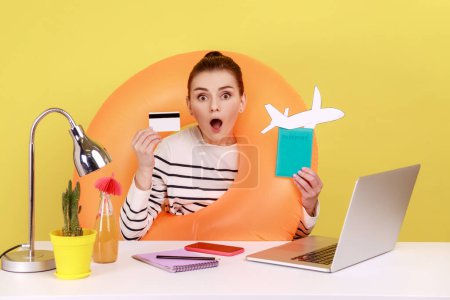 Photo for Amazed shocked woman with rubber ring holding and showing credit card and passport, booking tickets and hotel online on laptop. Indoor studio studio shot isolated on yellow background. - Royalty Free Image