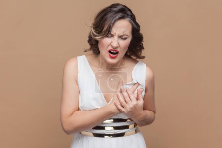 Photo for Middle aged woman with wavy hair holding hands on chest feeling acute pain in chest, risk of stroke, heart attack, wearing white dress. Indoor studio shot isolated on light brown background. - Royalty Free Image