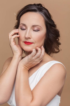 Photo for Closeup portrait of calm attractive beautiful middle aged woman with wavy hair and makeup, keeps eyes closed, wearing white dress. Indoor studio shot isolated on light brown background. - Royalty Free Image