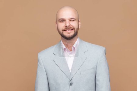 Photo for Portrait of playful positive optimistic bald bearded man has fun and winks eye, smiles with teeth makes funny grimace, wearing gray jacket. Indoor studio shot isolated on brown background. - Royalty Free Image
