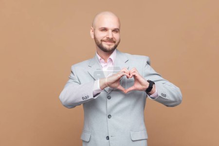 Photo for Portrait of bald bearded man shapes heart over chest, confesses in love, has romantic mood, expresses truthful feelings, wearing gray jacket. Indoor studio shot isolated on brown background. - Royalty Free Image