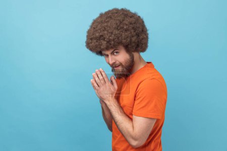 Photo for Portrait of man with Afro hairstyle wearing orange T-shirt rubbing palm as having cunning evil idea, devious plan in mind, thinking revenge. Indoor studio shot isolated on blue background. - Royalty Free Image