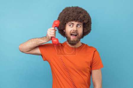 Photo for Portrait of man with Afro hairstyle wearing orange T-shirt talking landline telephone holding in hand handset, looking away with excited expression. Indoor studio shot isolated on blue background. - Royalty Free Image
