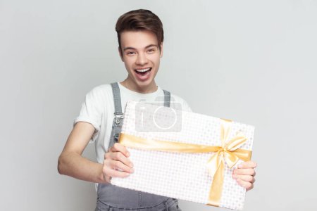 Photo for Portrait of joyful attractive excited brunette man standing standing with present box in hands, congratulating with holiday, wearing denim overalls. Indoor studio shot isolated on gray background. - Royalty Free Image