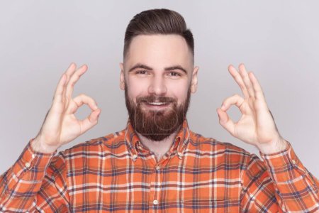 Photo for Smiling cheerful joyful bearded man standing looking at camera, showing okay sign, ok gesture, expressing positive emotions, wearing checkered shirt. Indoor studio shot isolated on gray background. - Royalty Free Image