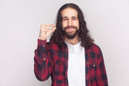 Photo for Bearded man with long curly hair in checkered red shirt shows something very small or tiny, discusses prices in store, smiles happily, says little bit. Indoor studio shot isolated on gray background. - Royalty Free Image