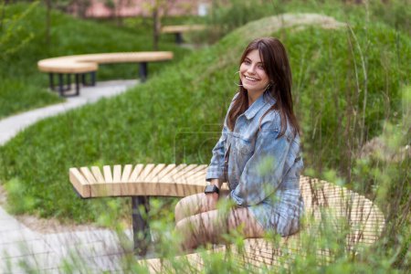 Téléchargez les photos : Portrait latéral d'une femme extrêmement heureuse portant une veste en denim, se reposant seule en plein air, profitant d'une chaude journée d'été, regardant la caméra sourire, assise sur un banc en bois dans un parc verdoyant. Plan extérieur. - en image libre de droit