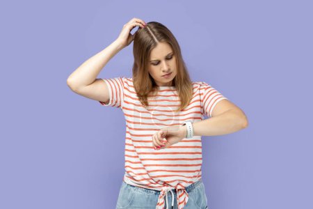 Photo for Portrait of pensive thoughtful blond woman wearing striped T-shirt standing looking at her smart watch and rubbing her head, thinking about deadline. Indoor studio shot isolated on purple background. - Royalty Free Image