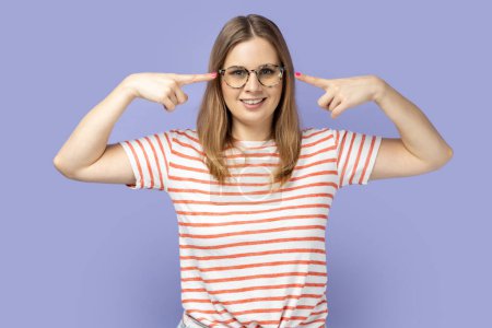 Photo for Portrait of delighted young adult blond woman wearing striped T-shirt standing and pointing at her spectacles, looking at camera with optimistic look. Indoor studio shot isolated on purple background. - Royalty Free Image