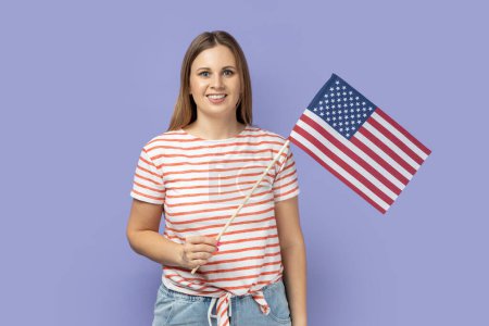 Photo for Portrait of good looking blond woman wearing striped T-shirt holding USA national flag, celebrating national Independence Day - 4th july. Indoor studio shot isolated on purple background. - Royalty Free Image