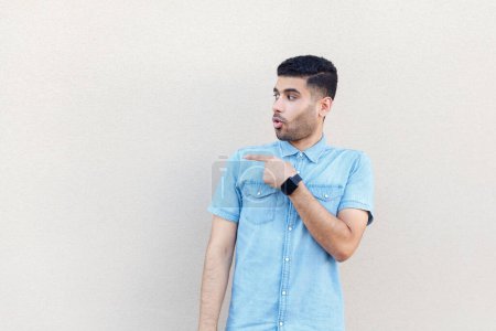Photo for Portrait of excited astonished man wearing denim shirt looking and pointing away at advertisement area, copy space for promotion. Indoor studio shot isolated on gray background. - Royalty Free Image