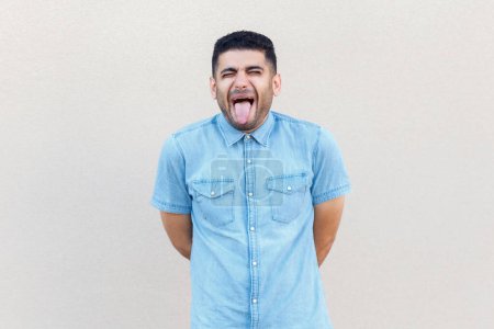 Photo for Portrait of funny crazy handsome man wearing denim shirt standing with closed eyes and showing tongue out, demonstrates childish behavior. Indoor studio shot isolated on gray background. - Royalty Free Image