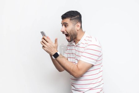 Photo for Side view portrait of amazed excited handsome bearded man wearing striped t-shirt standing using cell phone, reading shocking news. Indoor studio shot isolated on gray background. - Royalty Free Image