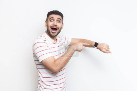 Photo for Portrait of amazed positive optimistic impressed bearded man wearing striped t-shirt standing looking at camera, pointing at smart watch. Indoor studio shot isolated on gray background. - Royalty Free Image