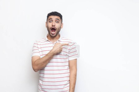 Photo for Portrait of astonished shocked bearded man wearing striped t-shirt standing pointing aside at copy space for promotion, advertisement area. Indoor studio shot isolated on gray background. - Royalty Free Image
