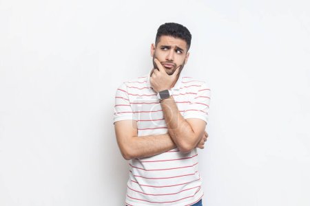 Photo for Portrait of pensive thoughtful attractive young adult bearded man wearing striped t-shirt standing holding chin, pondering, thinking. Indoor studio shot isolated on gray background. - Royalty Free Image