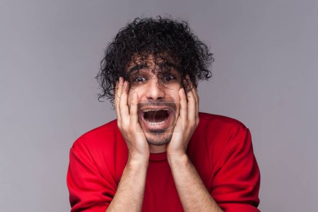 Photo for Portrait of shocked amazed bearded man with curly hair, looking in mirror, shocked from his hairstyle, keeps palms on cheeks, wearing red jumper. Indoor studio shot isolated on gray background. - Royalty Free Image