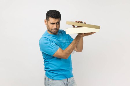 Photo for Portrait of attractive curious unshaven man wearing blue T- shirt standing holding gift box, looking inside, being interested what he received. Indoor studio shot isolated on gray background. - Royalty Free Image