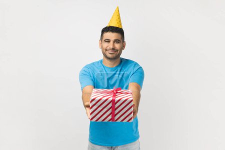 Photo for Portrait of cute attractive positive unshaven man wearing blue T- shirt and yellow party cone giving present box, having fun at birthday party. Indoor studio shot isolated on gray background. - Royalty Free Image