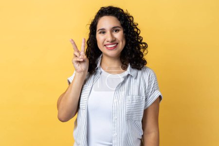 Photo for Portrait of happy optimistic woman with dark wavy hair doing victory gesture and looking smiling to camera, showing peace, v sign with double fingers. Indoor studio shot isolated on yellow background. - Royalty Free Image