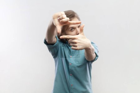 Photo for Portrait of man looking through frame of fingers gesturing photo, zooming focusing to camera, observing with serious expression, wearing blue shirt. Indoor studio shot isolated on gray background. - Royalty Free Image