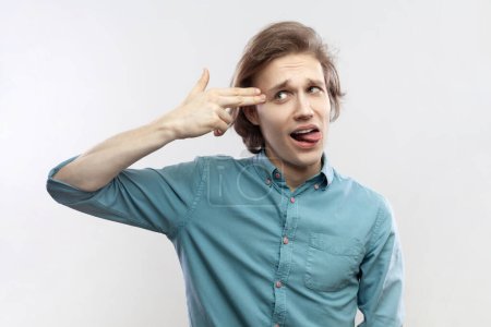 Photo for Portrait of depressed stressed handsome young man making suicide gesture, showing tongue out, having problems, wearing blue shirt. Indoor studio shot isolated on gray background. - Royalty Free Image