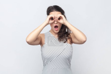 Photo for Portrait of surprised interested brunette woman making binoculars gesture with hands and looking amazed at camera, wearing striped dress. Indoor studio shot isolated on gray background. - Royalty Free Image
