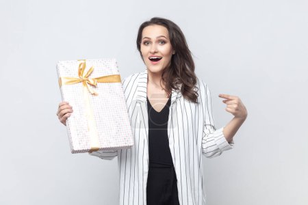 Photo for Portrait of surprised astonished brunette woman holding pointing gift box, looking at camera with big eyes and open mouth, wearing striped jacket. Indoor studio shot isolated on gray background. - Royalty Free Image