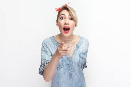Photo for Portrait of surprised shocked blonde woman wearing blue denim shirt and red headband standing, pointing at you, looking with big eyes. Indoor studio shot isolated on gray background. - Royalty Free Image
