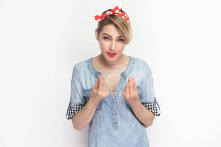Photo for Portrait of attractive beautiful blonde woman wearing blue denim shirt and red headband standing showing money gesture with fingers. Indoor studio shot isolated on gray background. - Royalty Free Image