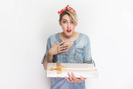 Photo for Portrait of surprised blonde woman wearing blue denim shirt and red headband standing holding present box, gets unexpected gift. Indoor studio shot isolated on gray background. - Royalty Free Image