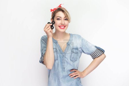 Photo for Portrait of smiling cheerful joyful blonde woman wearing blue denim shirt and red headband standing holding spinner, keeps hand on hips. Indoor studio shot isolated on gray background. - Royalty Free Image