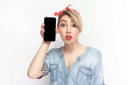 Téléchargez les photos : Portrait d'une jeune femme blonde choquée portant une chemise en denim bleu et un bandeau rouge tenant un téléphone intelligent avec écran blanc noir. Studio intérieur tourné isolé sur fond gris. - en image libre de droit