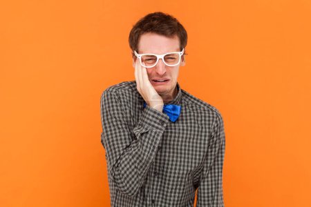 Man nerd with sorrowful expression, cries with despair, regrets doing something wrong, squints face, wearing shirt with blue bow tie and white glasses. Indoor studio shot isolated on orange background