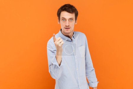 Photo for Portrait of serious bossy man standing looking at camera, raising his finger up, warning you about danger, wearing light blue shirt. Indoor studio shot isolated on orange background. - Royalty Free Image