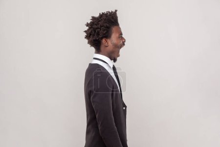 Photo for Side view portrait of man shouting loudly having aggression, screaming with wide opened mouth, having unhappy expression, wearing white shirt and tuxedo. Indoor studio shot isolated on gray background - Royalty Free Image