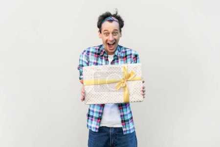 Photo for Excited man giving present box to his friend, being in good festive mood, screaming in astonishment, wearing blue checkered shirt and headband. Indoor studio shot isolated on gray background. - Royalty Free Image