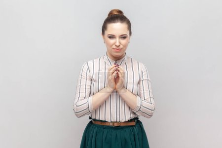 Photo for Portrait of cunning young adult woman wearing striped shirt and green skirt keeps hands together, having slander plan, pondering. Indoor studio shot isolated on gray background. - Royalty Free Image