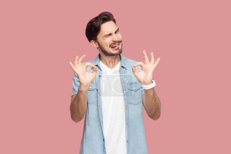 Photo for Its excellent result. Portrait of bearded man in blue casual style shirt makes okay gesture, enjoys life, says ok, confirms information. Indoor studio shot isolated on pink background. - Royalty Free Image