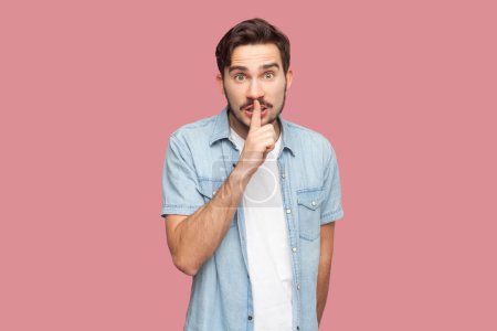 Photo for Portrait of stupefied secret bearded man in blue casual style shirt standing makes gesture quietly, asks remain silent, gossips about something. Indoor studio shot isolated on pink background. - Royalty Free Image