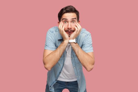 Photo for Portrait of shocked astonished bearded man in blue casual style shirt standing with hands on cheeks, looking at camera with big eyes. Indoor studio shot isolated on pink background. - Royalty Free Image